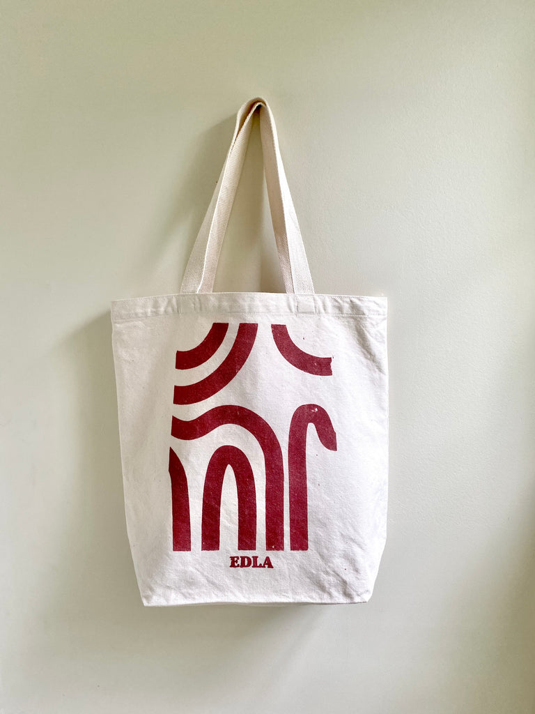 White cotton tote bag against white wall. Tote bag has red design made of curved lines and text in same color at bottom which says Edla